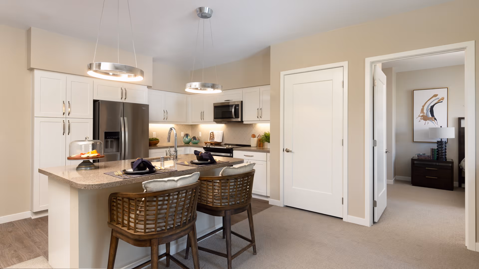 Modern kitchen with white cabinets, stainless steel refrigerator and microwave, an island with a sink and two wooden bar stools with cushions. The kitchen is well-lit with two circular pendant lights hanging above the island. A doorway leads to a bedroom with a nightstand, lamp, and abstract wall art.