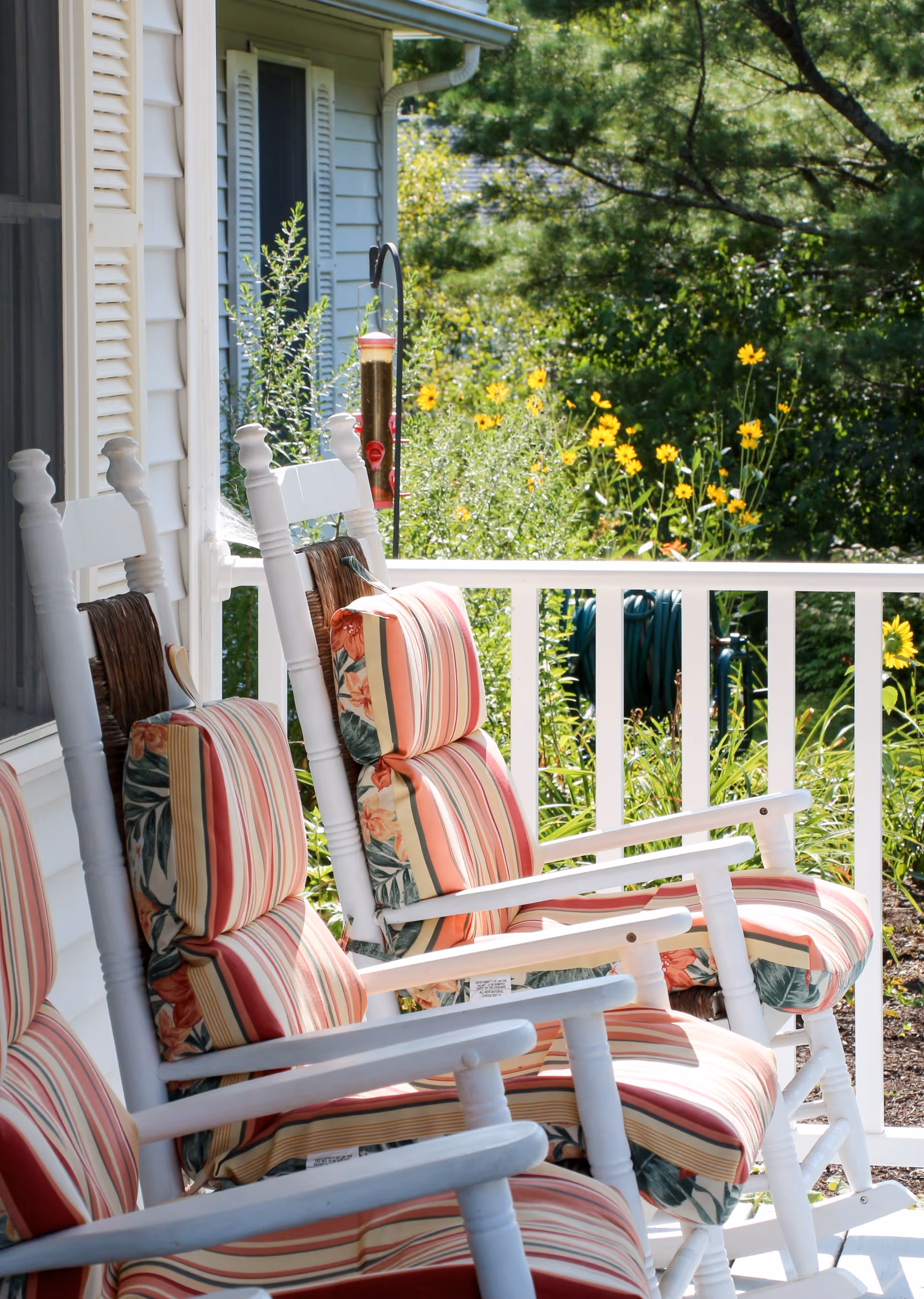 Three white wooden rocking chairs with striped and floral cushions on a porch railing, overlooking a garden with yellow flowers and green foliage in bright sunlight.