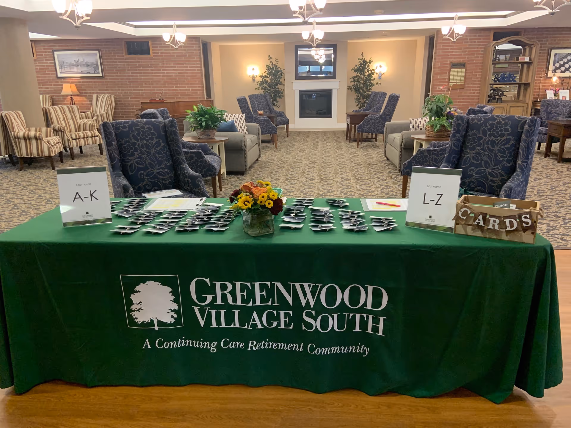 A greeting table covered with a green 'Greenwood Village South' tablecloth holding name-tag piles, signs, and a flower arrangement in a furnished community lounge.