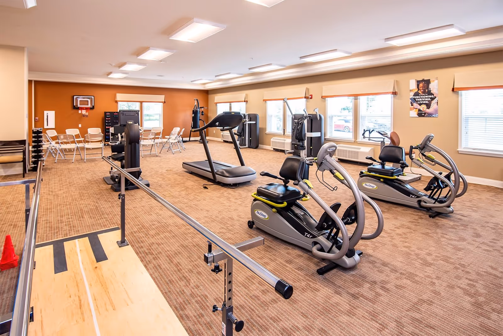 A spacious fitness room in a senior living facility with exercise equipment including two NuStep recumbent cross trainers, a treadmill, and strength training machines. The room has beige walls, multiple windows with beige valances, and a carpeted floor. There are parallel bars for walking exercises and several white folding chairs arranged near a small basketball hoop on the far wall. A motivational poster is visible on the wall.