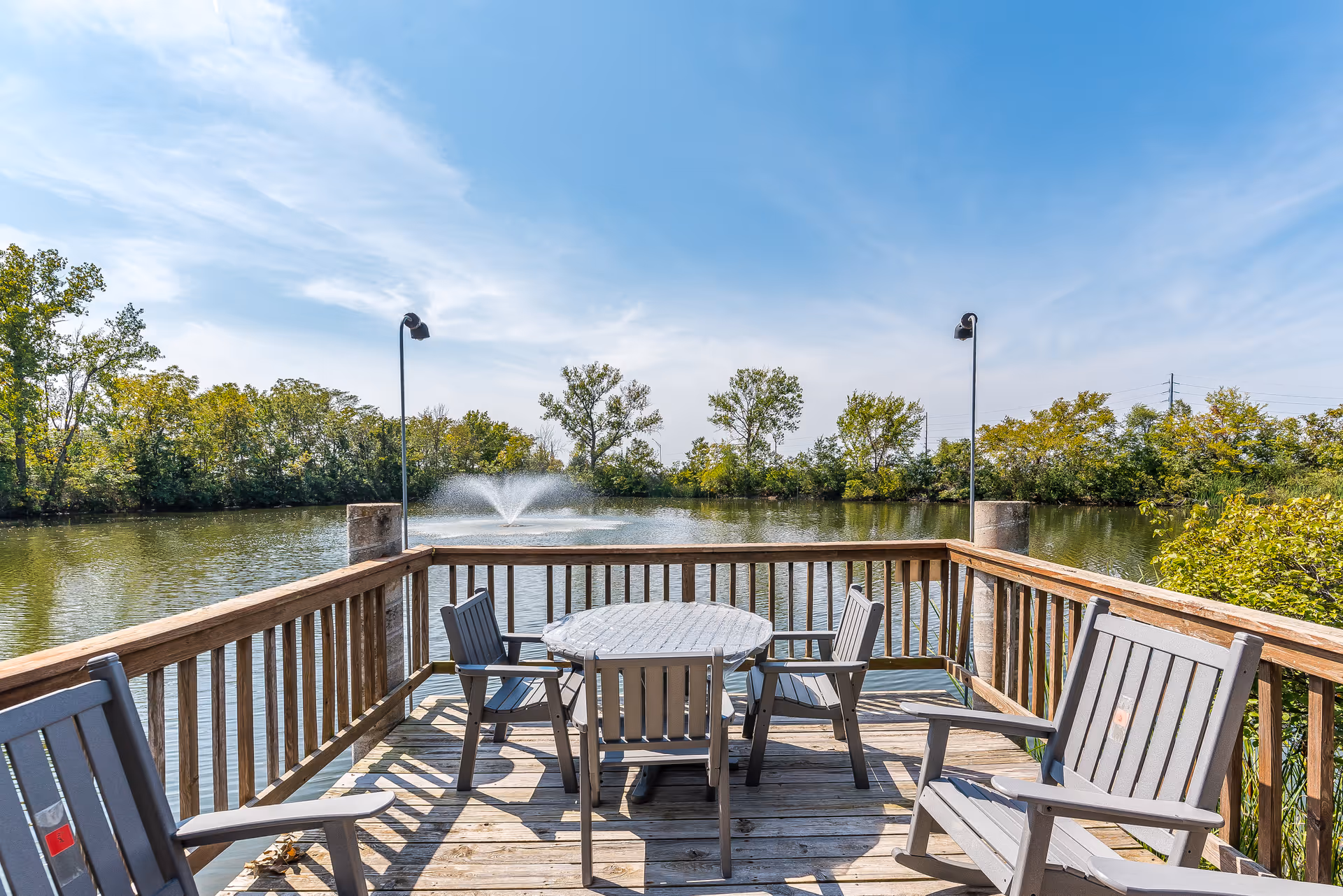 Wooden deck with a round table and chairs overlooking a pond with a fountain and trees under a blue sky.
