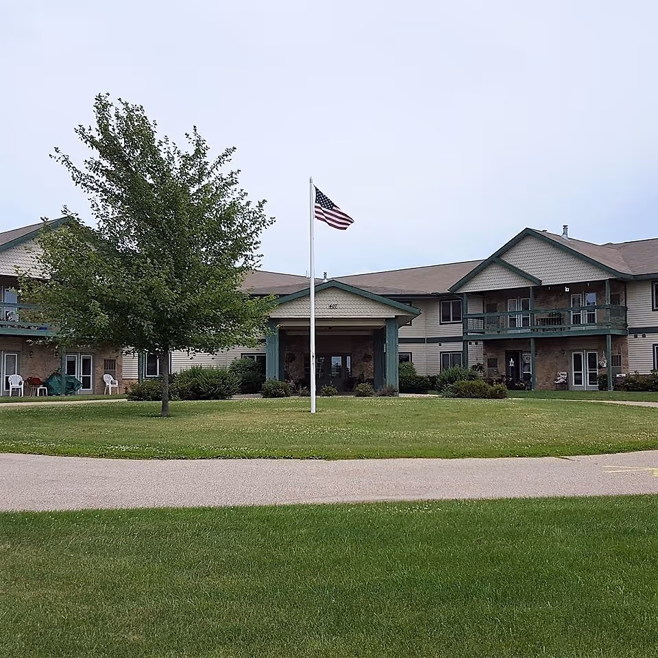 Exterior view of Stonefield Apartment Homes & Health Services building with a large green lawn, a tree, and an American flag on a flagpole in front of the entrance.