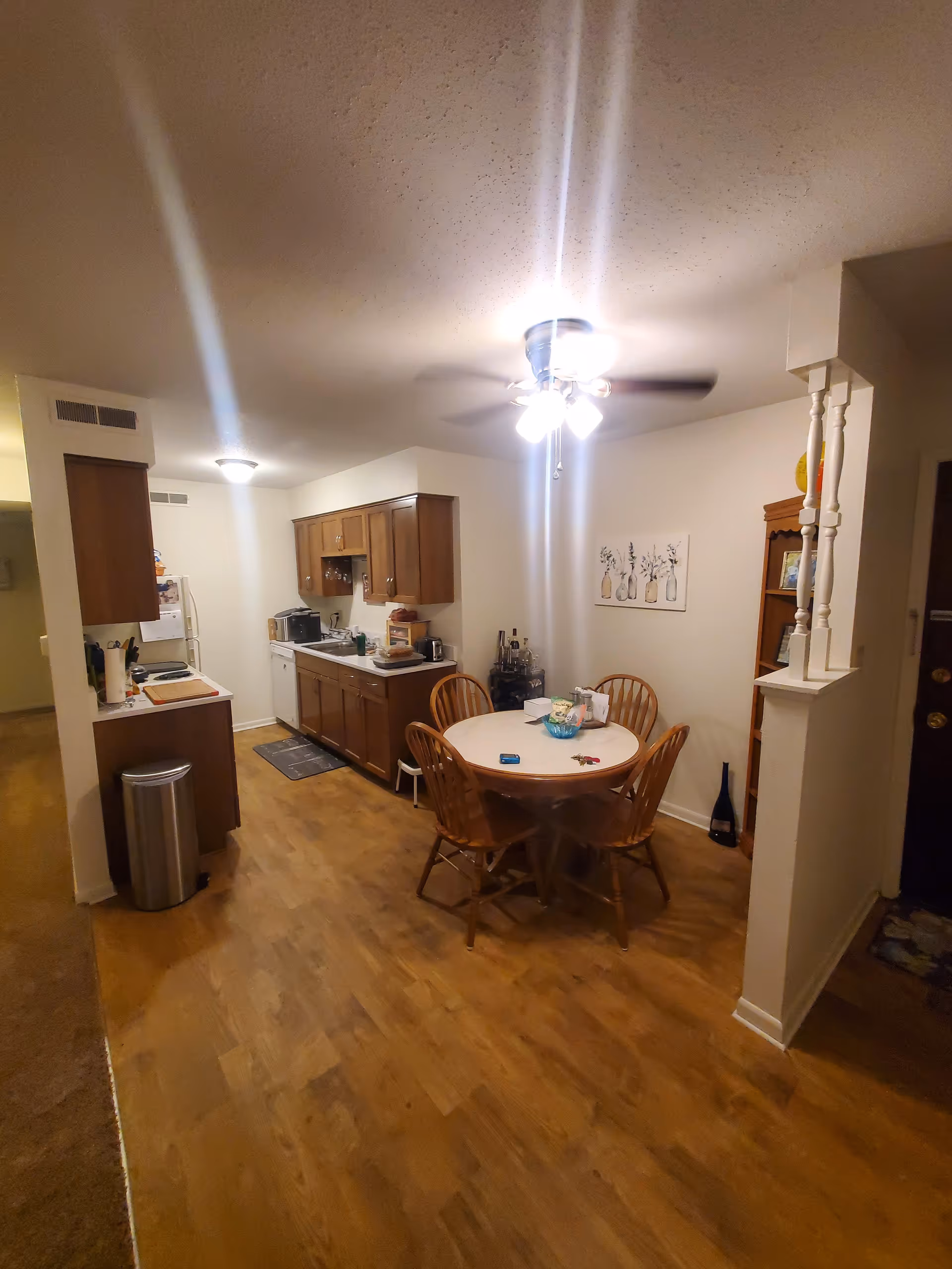 Interior view of a kitchen and dining area in an apartment. The kitchen has wooden cabinets, a white refrigerator, a dishwasher, and a countertop with various kitchen items. The dining area features a round wooden table with four matching chairs, a ceiling fan with lights above, and a wooden shelving unit against the wall. The floor is wood laminate, and the walls are painted white.