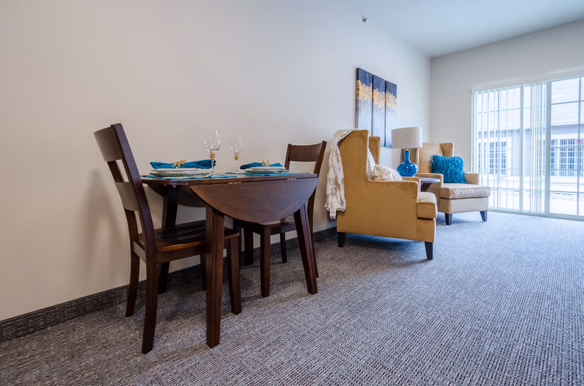 A cozy interior space featuring a small wooden dining table set with two chairs, two beige armchairs with a blue pillow and a white throw blanket, a blue table lamp on a side table, and a large window with vertical blinds allowing natural light to fill the room.