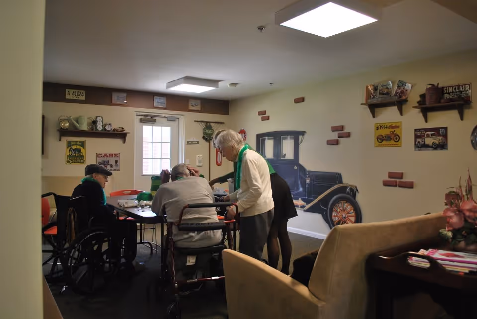 A group of elderly people gathered around a table in a cozy room decorated with vintage car-themed wall art and shelves holding various items. One person is in a wheelchair, another uses a walker, and a caregiver is assisting them. The room has a door with windows and soft lighting from ceiling fixtures.