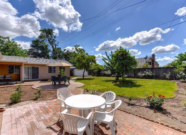 Outdoor patio area with a round white table and four white plastic chairs on a brick surface. In the background, there is a green lawn with trees and bushes, a beige single-story building with sliding glass doors, and a blue sky with scattered clouds.