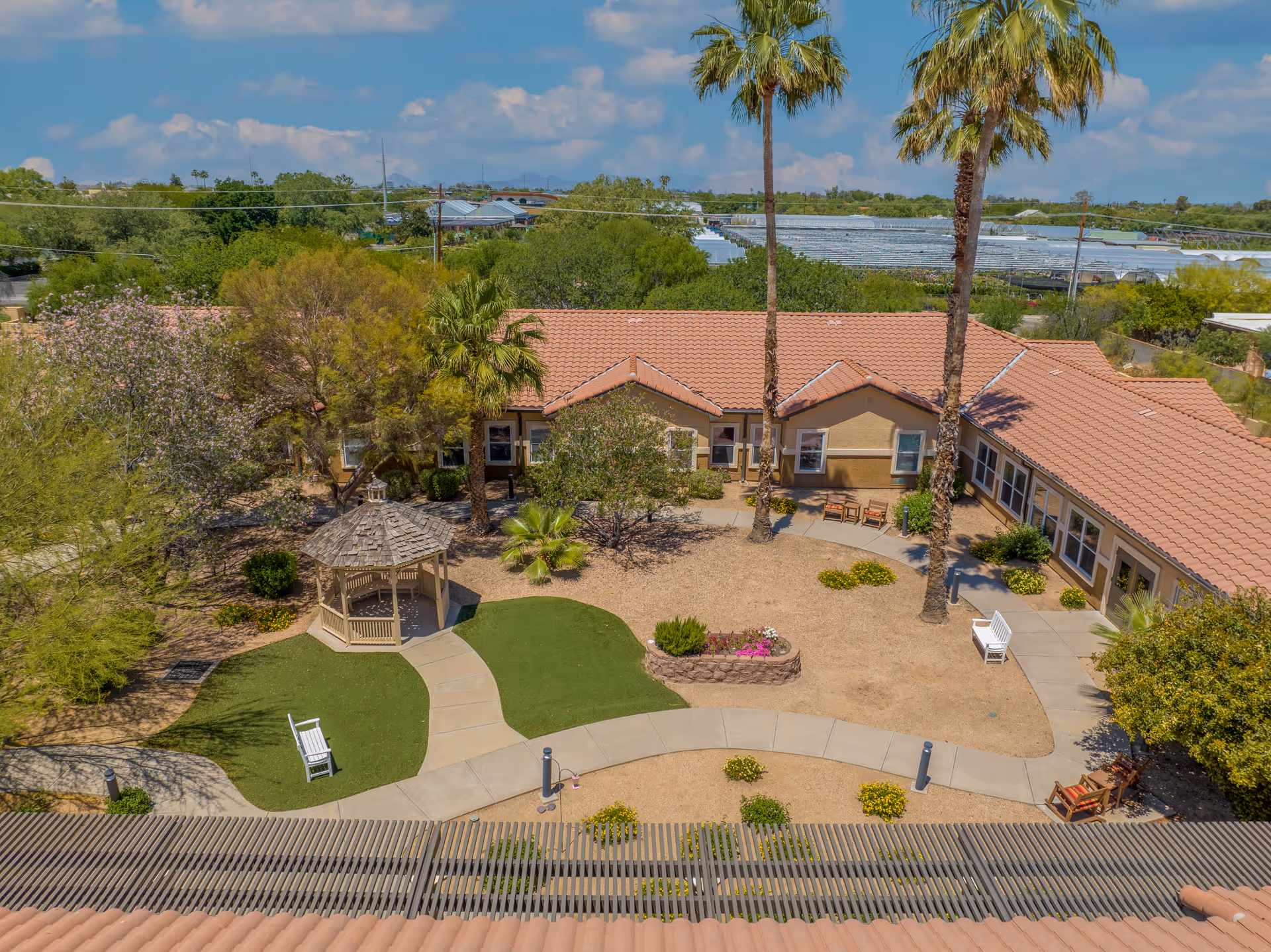 Aerial view of an assisted living facility courtyard with a gazebo, palm trees, benches, and landscaped garden areas surrounded by a single-story building with a tiled roof under a partly cloudy sky.