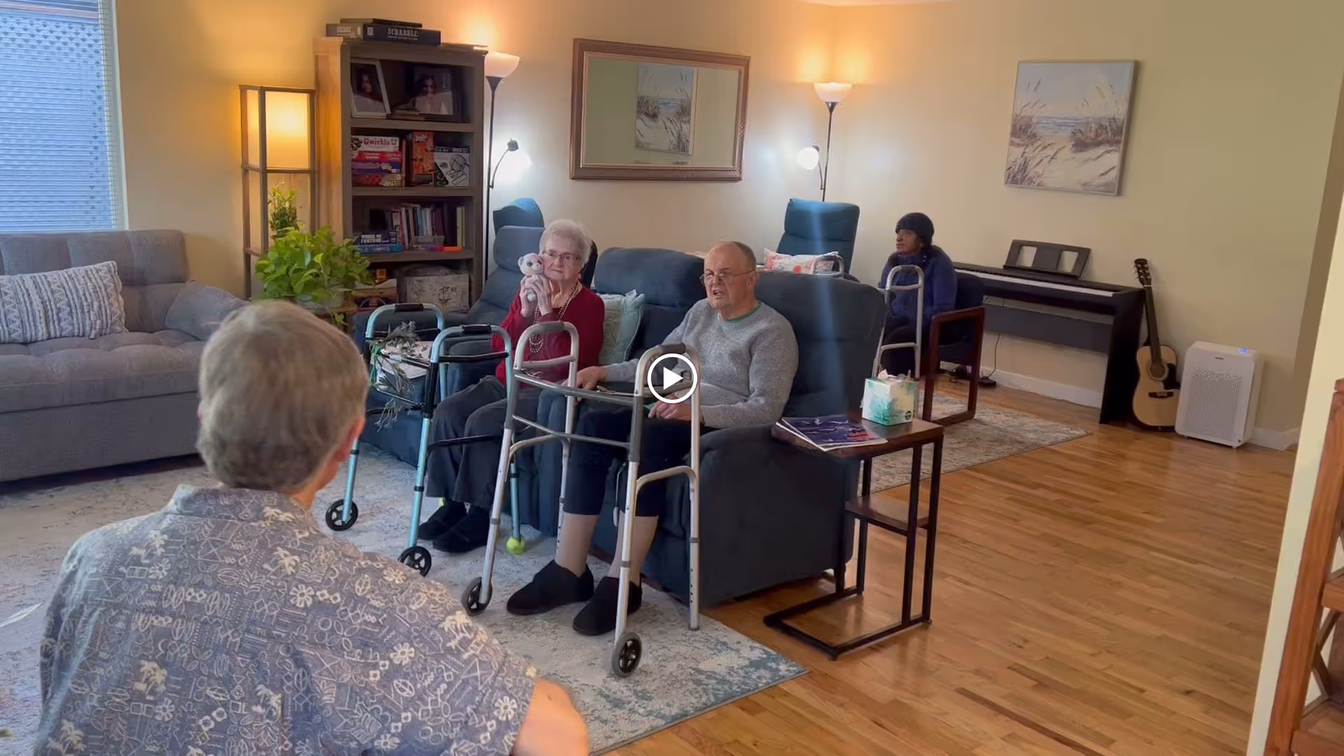 A cozy living room in an elderly care facility with three elderly individuals seated on armchairs and a sofa. Two of them have walkers in front of them. The room features wooden flooring, a bookshelf, a floor lamp, framed artwork on the walls, a piano with a guitar beside it, and a small side table with tissues.