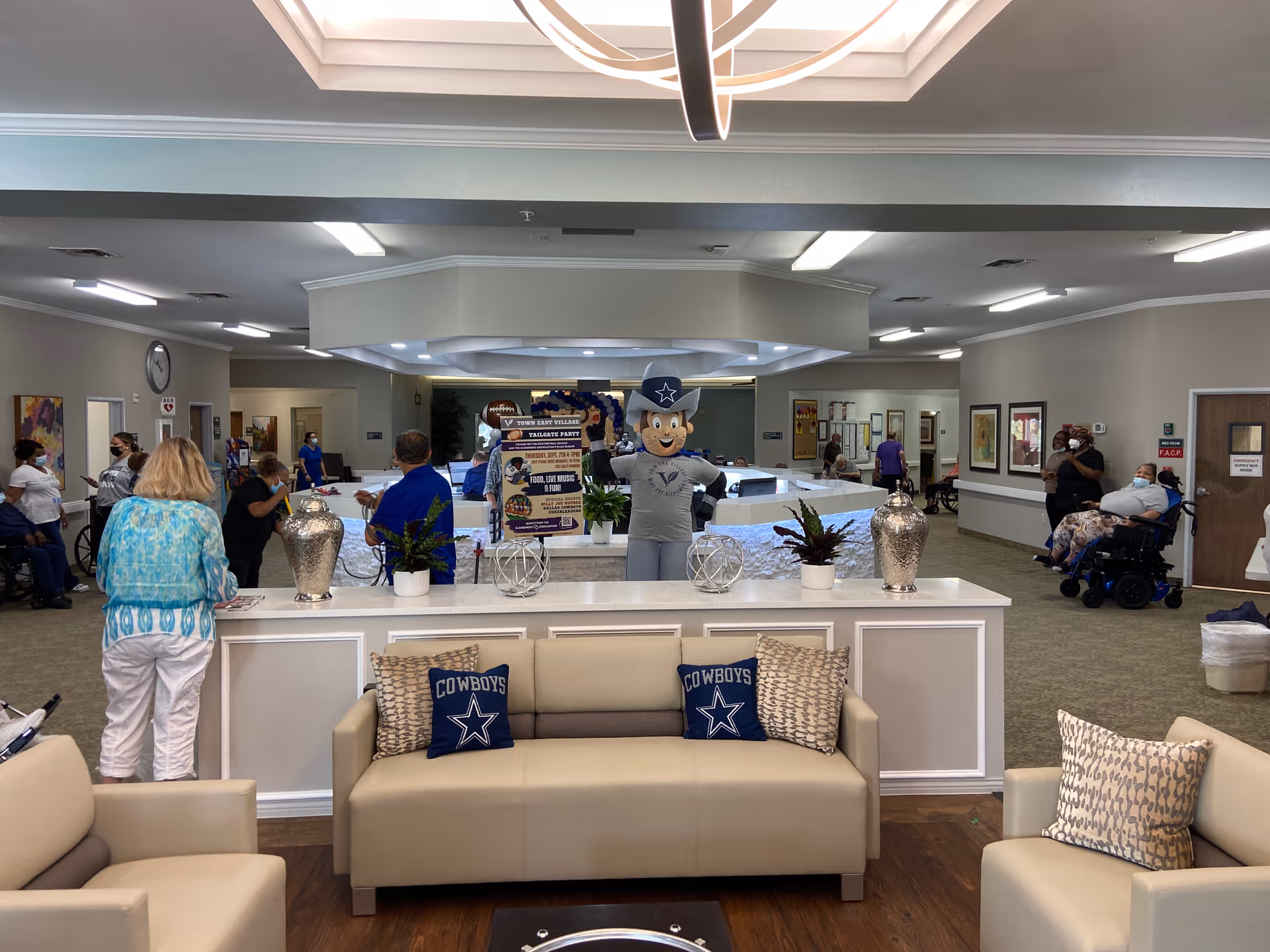 A senior living facility common area with beige couches and Cowboys-themed pillows in the foreground. Behind the couches is a white counter with decorative plants and vases. In the background, several people, some in wheelchairs and some standing, are gathered around a reception desk area. A mascot figure wearing a cowboy hat and a Town East Village shirt stands behind the counter holding a sign about a tailgate party event. The room has bright overhead lighting and neutral-colored walls with framed artwork.