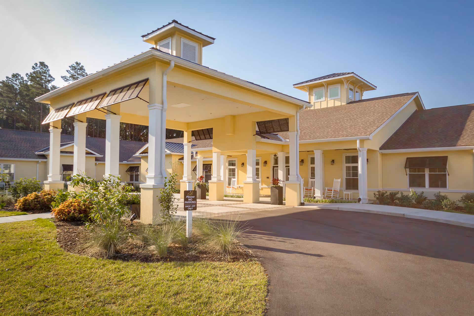 Exterior view of The Addison of Bluffton senior living facility entrance with a covered drop-off area, surrounded by landscaped greenery and a clear blue sky.