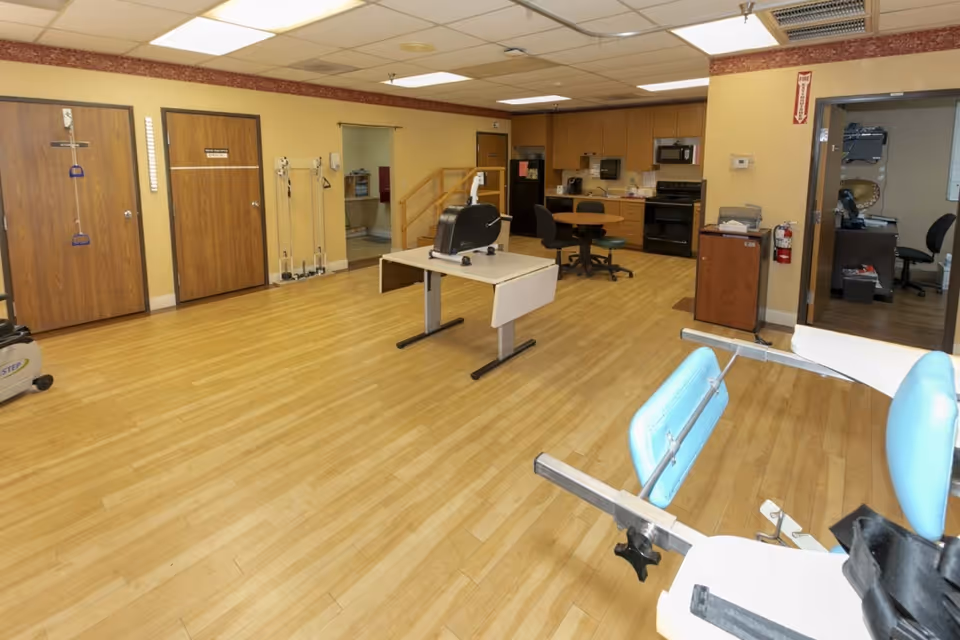 A spacious room with light wood flooring and beige walls, featuring exercise equipment including a stationary bike and a leg exercise machine. There are wooden doors on the left wall, a table with a black exercise machine in the center, and a small kitchen area with cabinets, a microwave, and a stove in the background. An office area with a desk and chair is visible through an open doorway on the right.