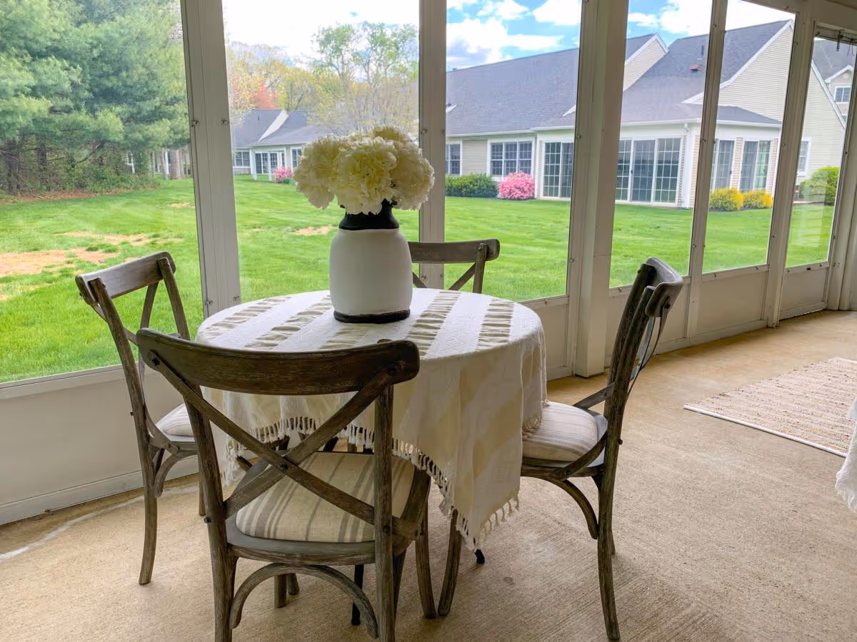 A small round table covered with a white tablecloth and a vase of white flowers on top, surrounded by four wooden chairs with cushions, situated in a sunroom with large windows overlooking a green lawn and neighboring buildings.
