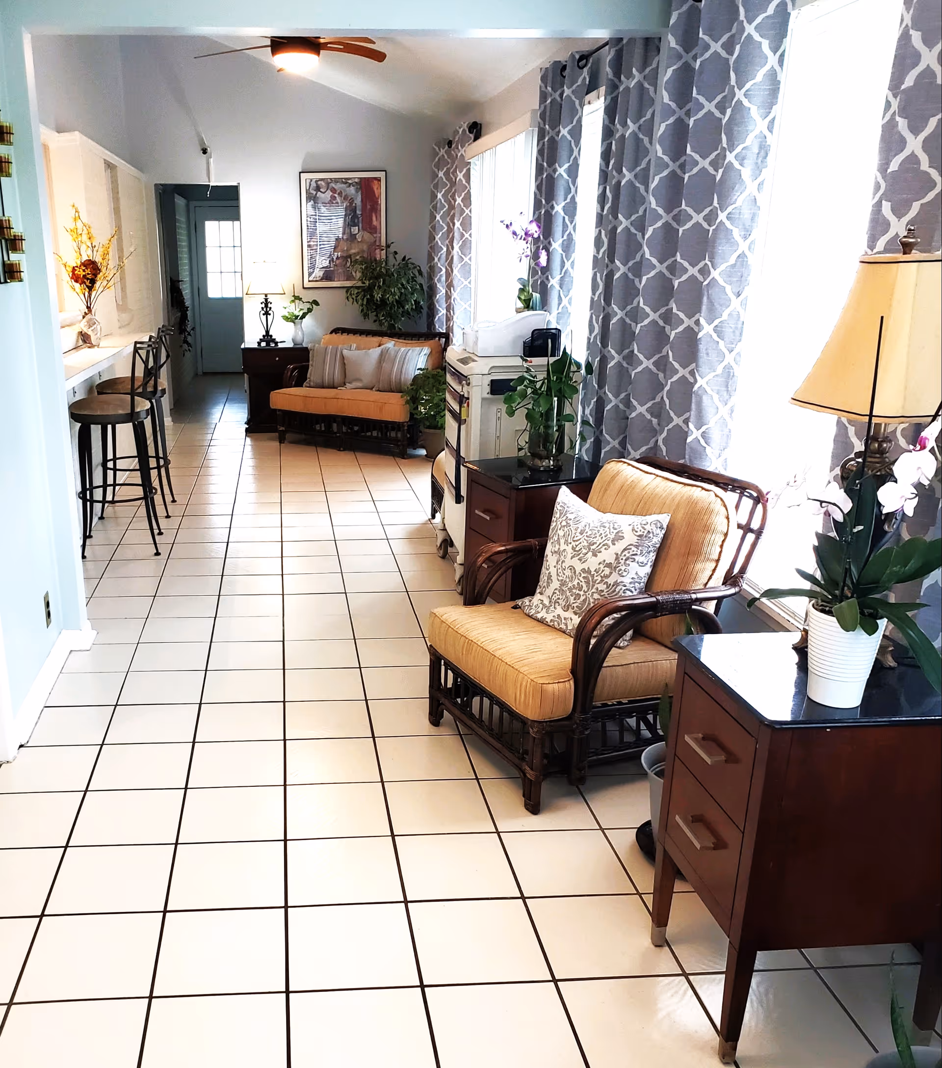 A bright, tiled hallway in a senior living facility with cushioned wicker chairs, side tables with lamps and potted plants, patterned curtains on large windows, and bar stools along a counter on the left side.