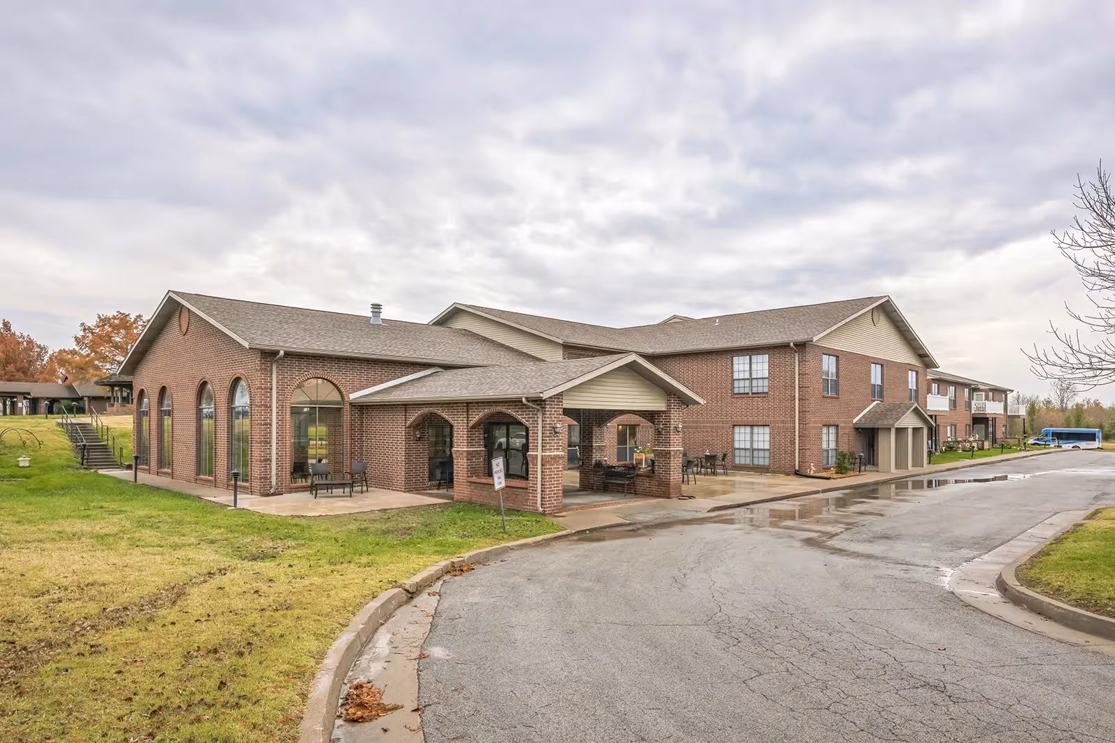 Exterior view of a two-story brick building with large arched windows and a covered entrance. The building is surrounded by a paved driveway and grassy areas with some trees. The sky is cloudy.