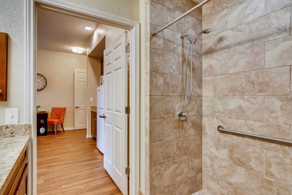 View of a bathroom shower area with beige and brown tiled walls, a silver showerhead, and a grab bar. The bathroom opens into a hallway with wood flooring, a white door, a stacked washer and dryer, and an orange chair against the wall with a clock above it.