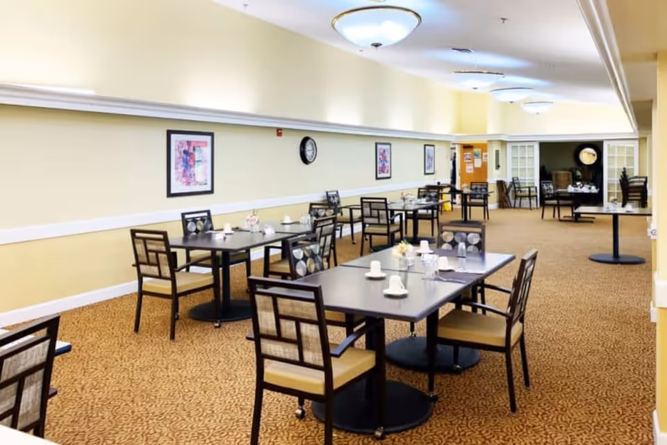 A spacious dining room in a senior living facility with multiple tables and chairs arranged neatly. The tables are set with cups, saucers, and small flower arrangements. The room has beige walls, carpeted floors with a patterned design, framed artwork on the walls, and ceiling lights providing bright illumination.