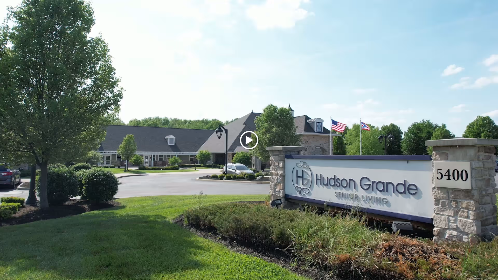 Exterior view of Hudson Grande Senior Living facility with a large sign displaying the facility name and address number 5400. The building is surrounded by trees, bushes, and a well-maintained lawn under a partly cloudy sky.