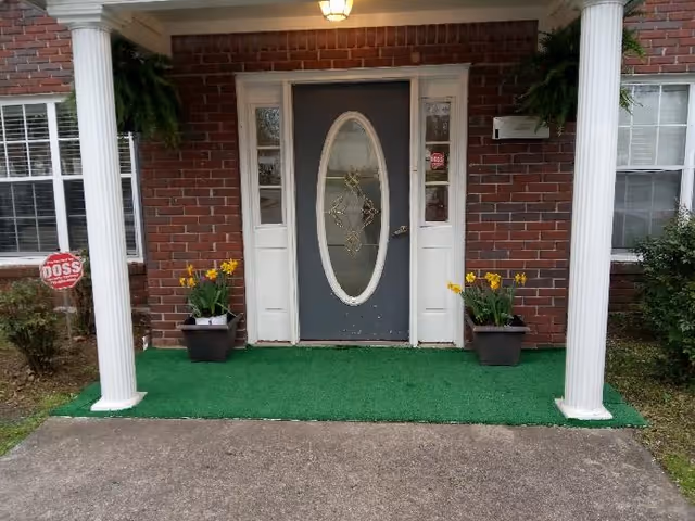 Front entrance of a brick building with a gray oval-glass door flanked by white columns, potted yellow flowers, hanging ferns and a green outdoor carpet.