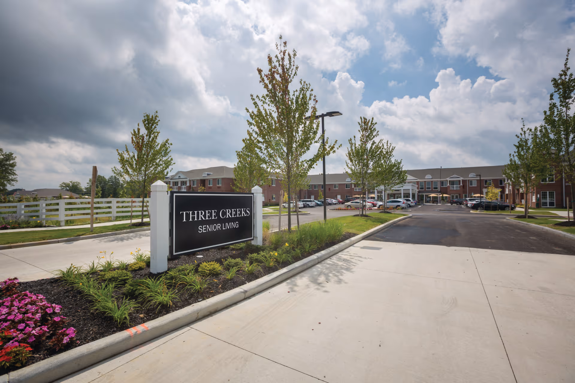Entrance driveway to a senior living facility with a landscaped area featuring green plants and purple flowers. A black sign with white text reads 'THREE CREEKS SENIOR LIVING'. The building is a two-story brick structure with multiple windows and a parking lot with several cars. The sky is partly cloudy.