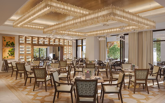 A spacious and elegant dining room with multiple wooden tables and chairs arranged neatly. The room features large windows with curtains allowing natural light to enter, decorative wall art, and a modern chandelier with a geometric design hanging from the ceiling. The floor has a patterned design, and the overall ambiance is warm and inviting.
