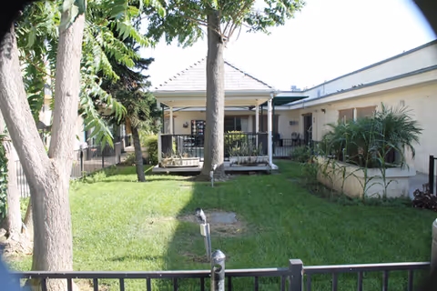 A grassy outdoor courtyard area with trees and a small covered patio attached to a single-story building. There is a black metal fence in the foreground and some plants along the building wall.
