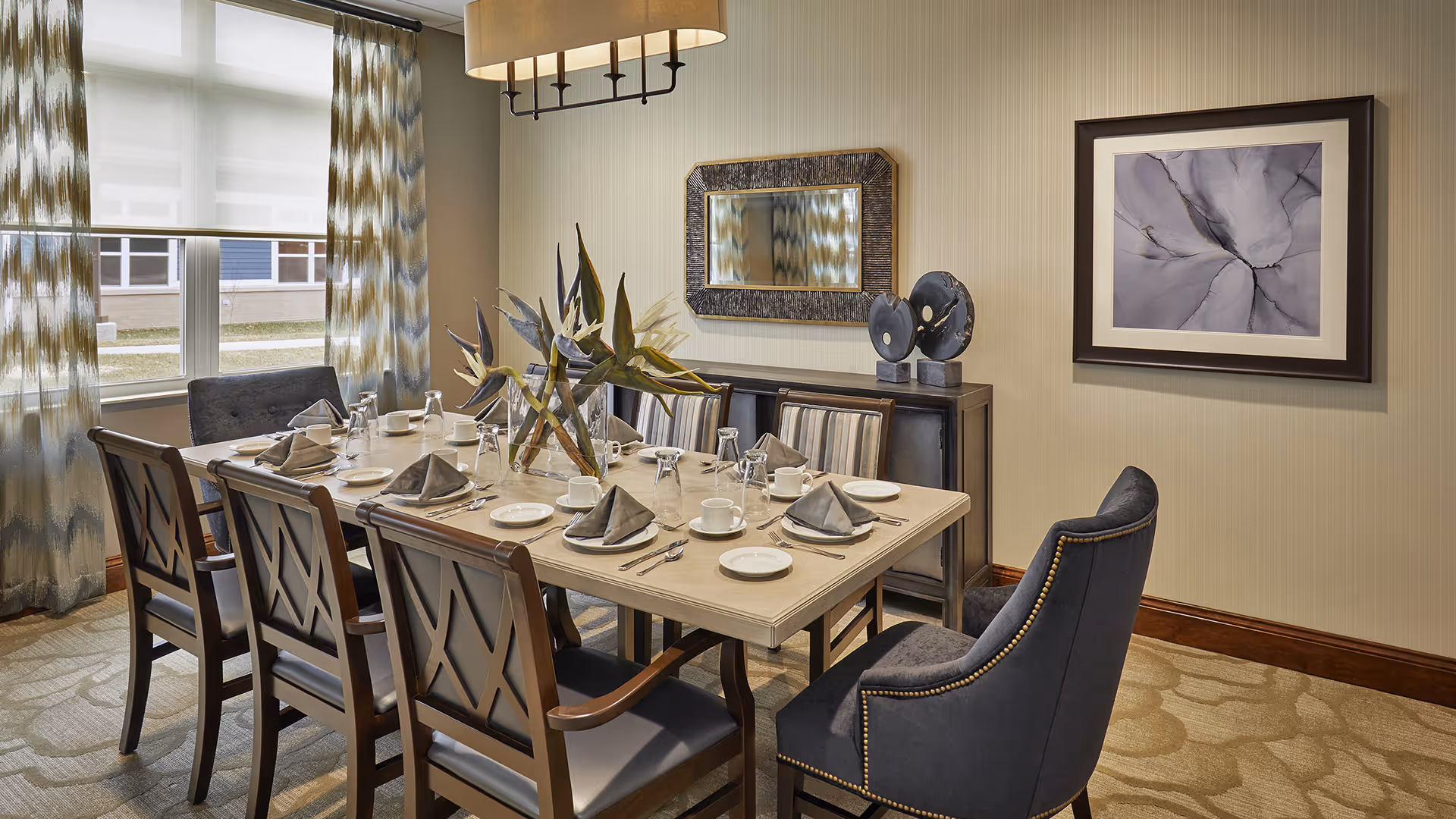 A well-lit dining room with a rectangular table set for eight people. The table is arranged with plates, cups, napkins, and silverware, and decorated with a centerpiece of bird of paradise flowers. The room features patterned curtains on the windows, a large mirror and decorative objects on a sideboard against the wall, and a framed abstract artwork. The chairs are a mix of wooden and upholstered styles, and a modern chandelier hangs above the table.