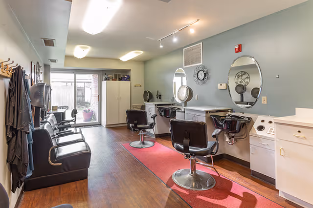 Interior view of a hair salon area in a senior living facility with salon chairs, mirrors, hair washing stations, and a seating area along the left wall. There is a red rug on the wooden floor and a sliding glass door at the far end leading outside.