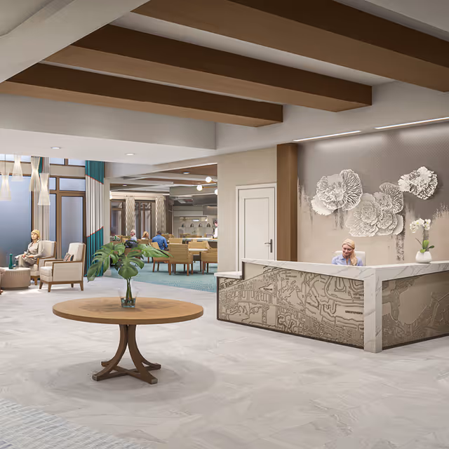 A spacious and modern senior living facility lobby with a round wooden table holding a vase with green leaves in the center. To the right, a receptionist sits behind a decorative front desk with artistic wall decor above. In the background, there are seating areas with chairs and people engaged in conversation, and large windows letting in natural light.