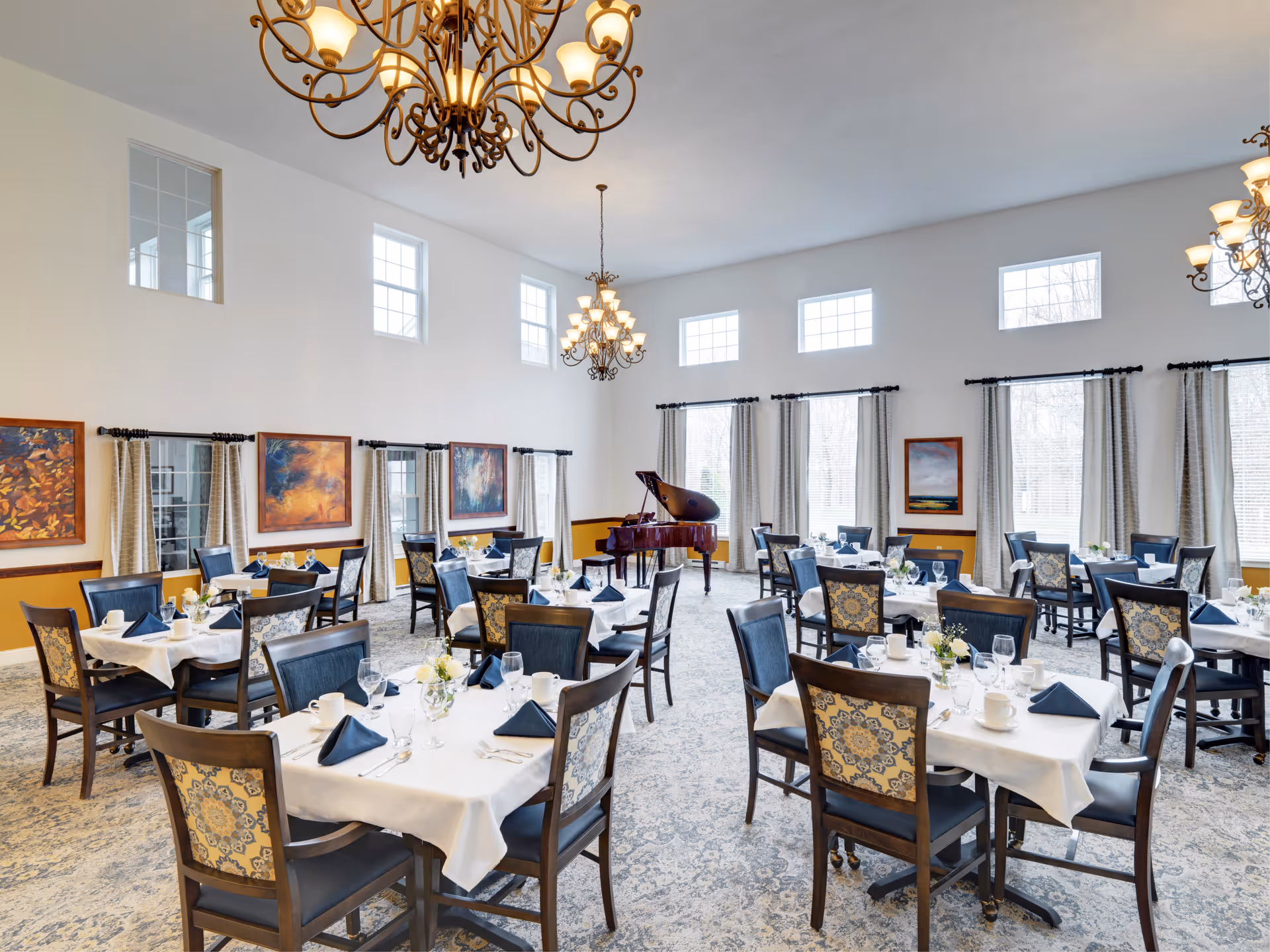 A bright and elegant dining room with multiple tables set with white tablecloths, navy blue napkins, glassware, and white cups. The room features large windows with curtains, several framed paintings on the walls, ornate chandeliers hanging from the ceiling, and a grand piano in the corner.