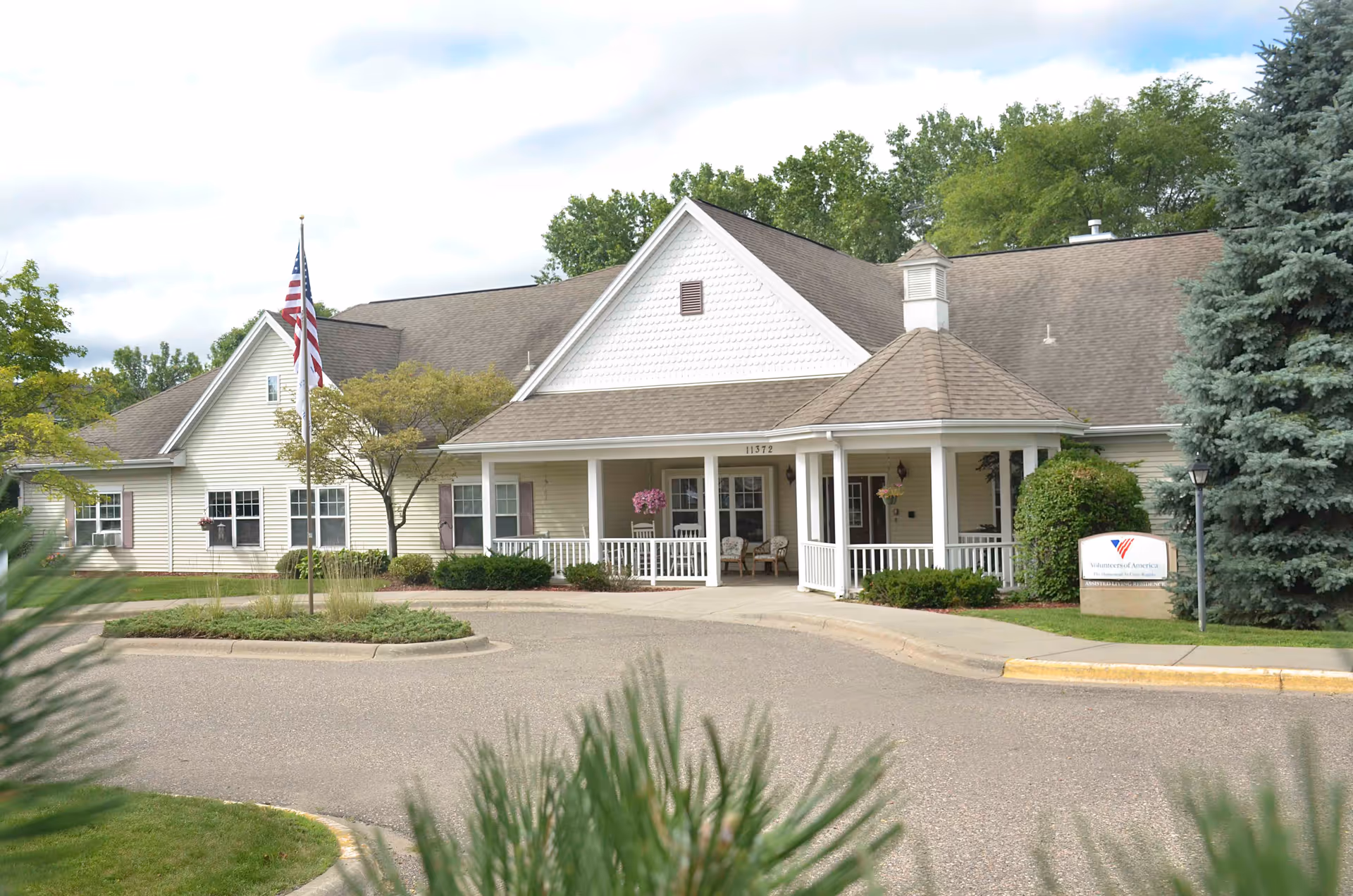 Exterior view of a single-story senior living facility building with a beige facade, white trim, and a covered porch with chairs. There is an American flag on a flagpole in front of the building, surrounded by trees and shrubs. A sign near the entrance reads Volunteers of America.