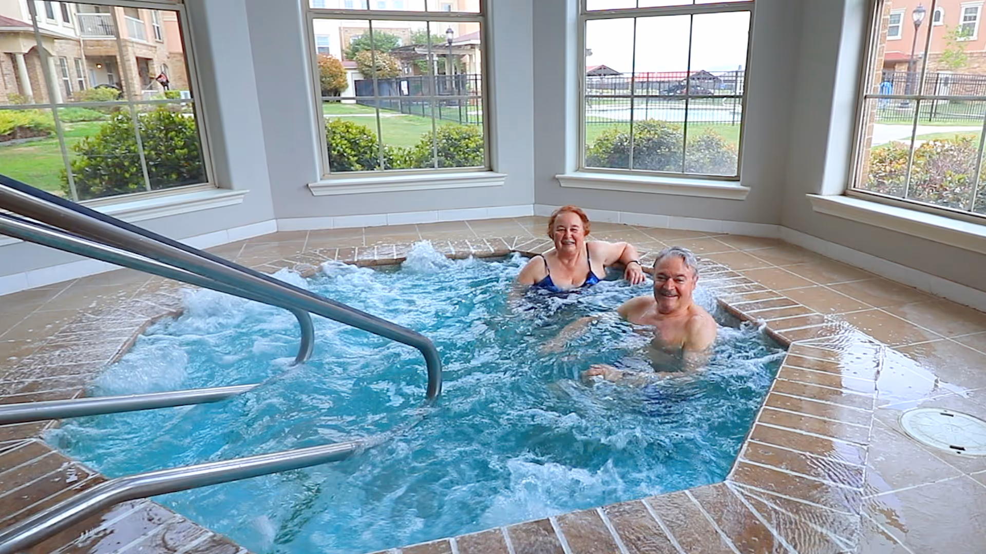 Two elderly people enjoying a bubbling indoor hot tub with large windows showing a green outdoor area and buildings in the background.