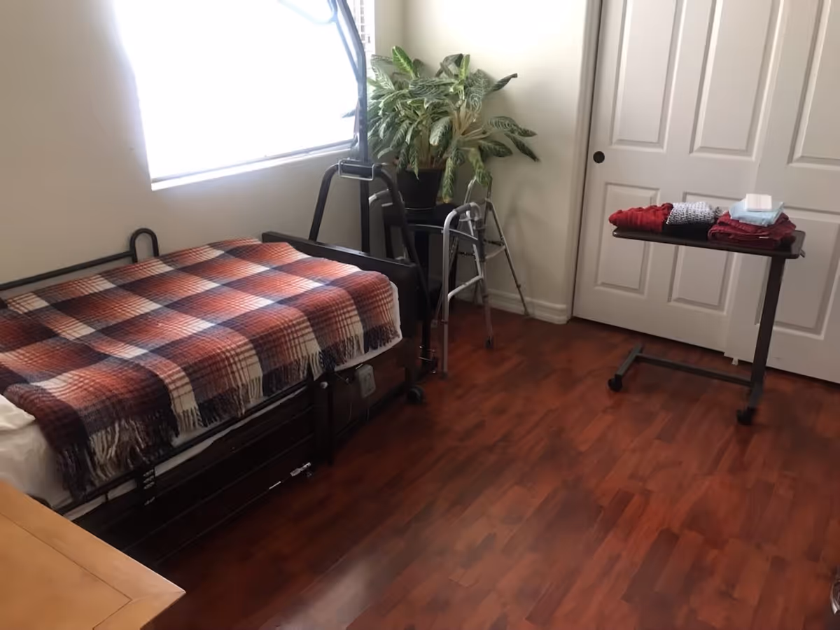 A small bedroom with a single bed covered by a red, white, and blue plaid blanket. Next to the bed is a walker and a potted plant on a stand. A small table with folded clothes and a pack of wipes is positioned near a closed white door. The room has wooden flooring and a window letting in natural light.