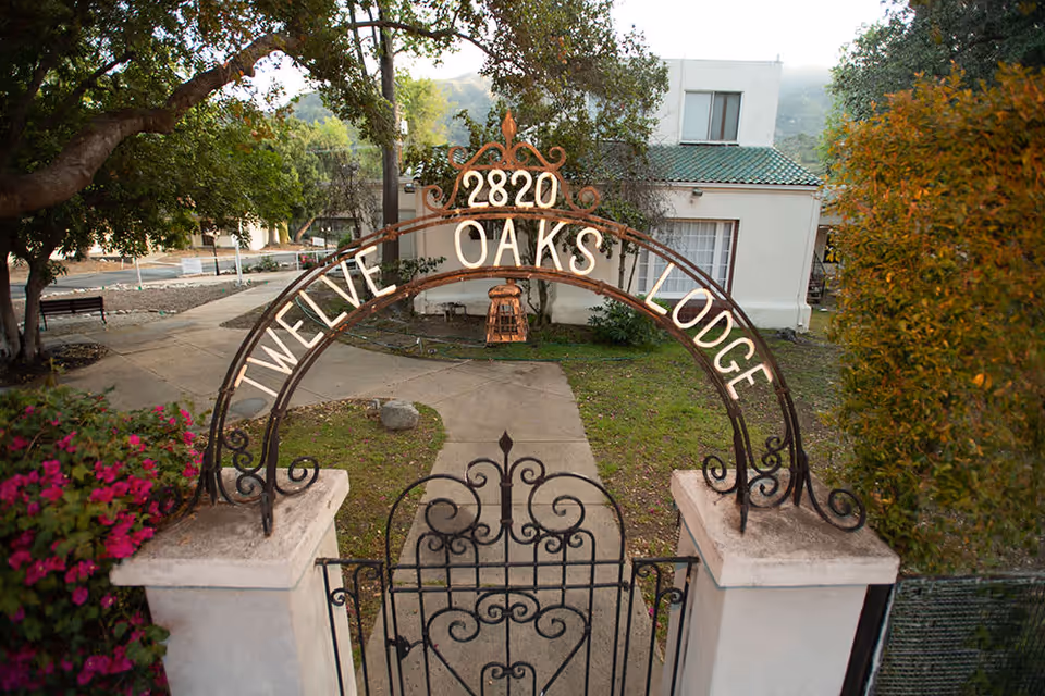 Entrance gate with an arch that reads '2820 Twelve Oaks Lodge' leading to a pathway surrounded by greenery and a white building with a green tiled roof in the background.