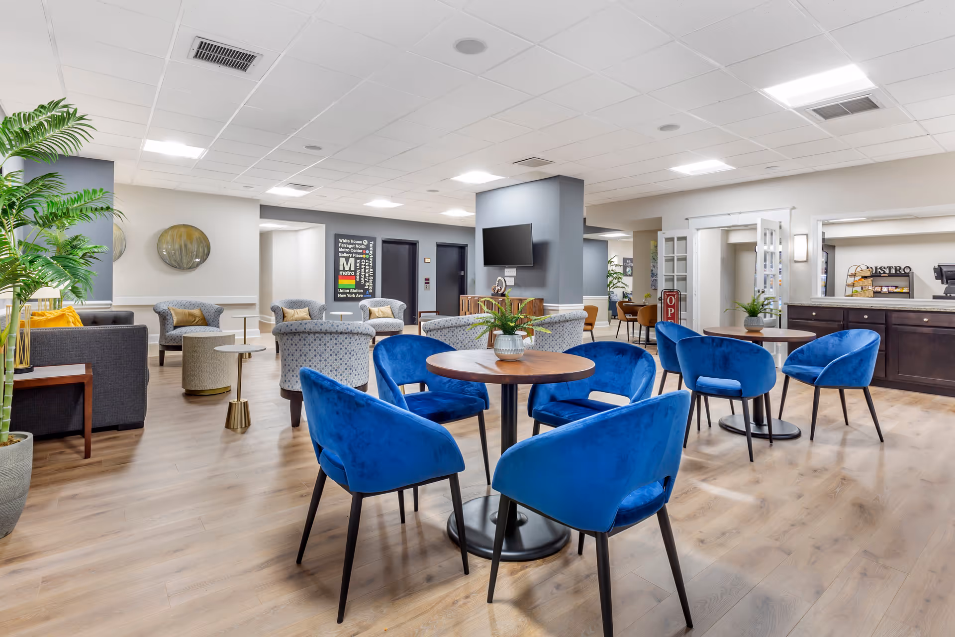 A spacious and well-lit common area in a retirement community featuring multiple seating arrangements. There are round wooden tables surrounded by blue upholstered chairs in the foreground. In the background, there are gray and patterned armchairs, a flat-screen TV mounted on a gray pillar, and a counter area with a coffee station. The room has light wood flooring, white walls, and a ceiling with recessed lighting. A large green plant is visible on the left side.
