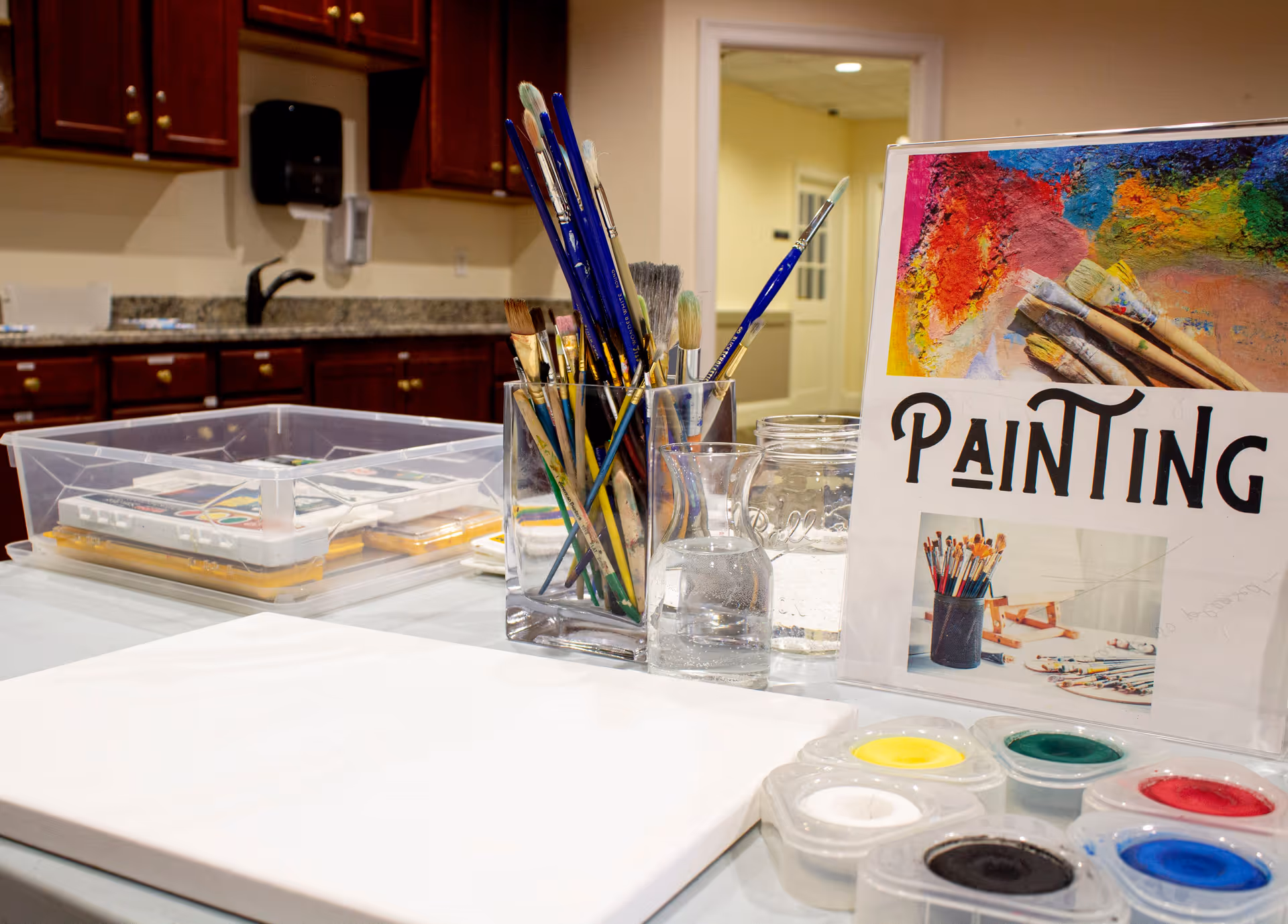 A table set up for painting with various paintbrushes in a clear container, small paint pots with different colors, a blank canvas, and a sign that reads 'PAINTING' with colorful artwork and paintbrushes pictured. The background shows a kitchen area with wooden cabinets and a granite countertop.