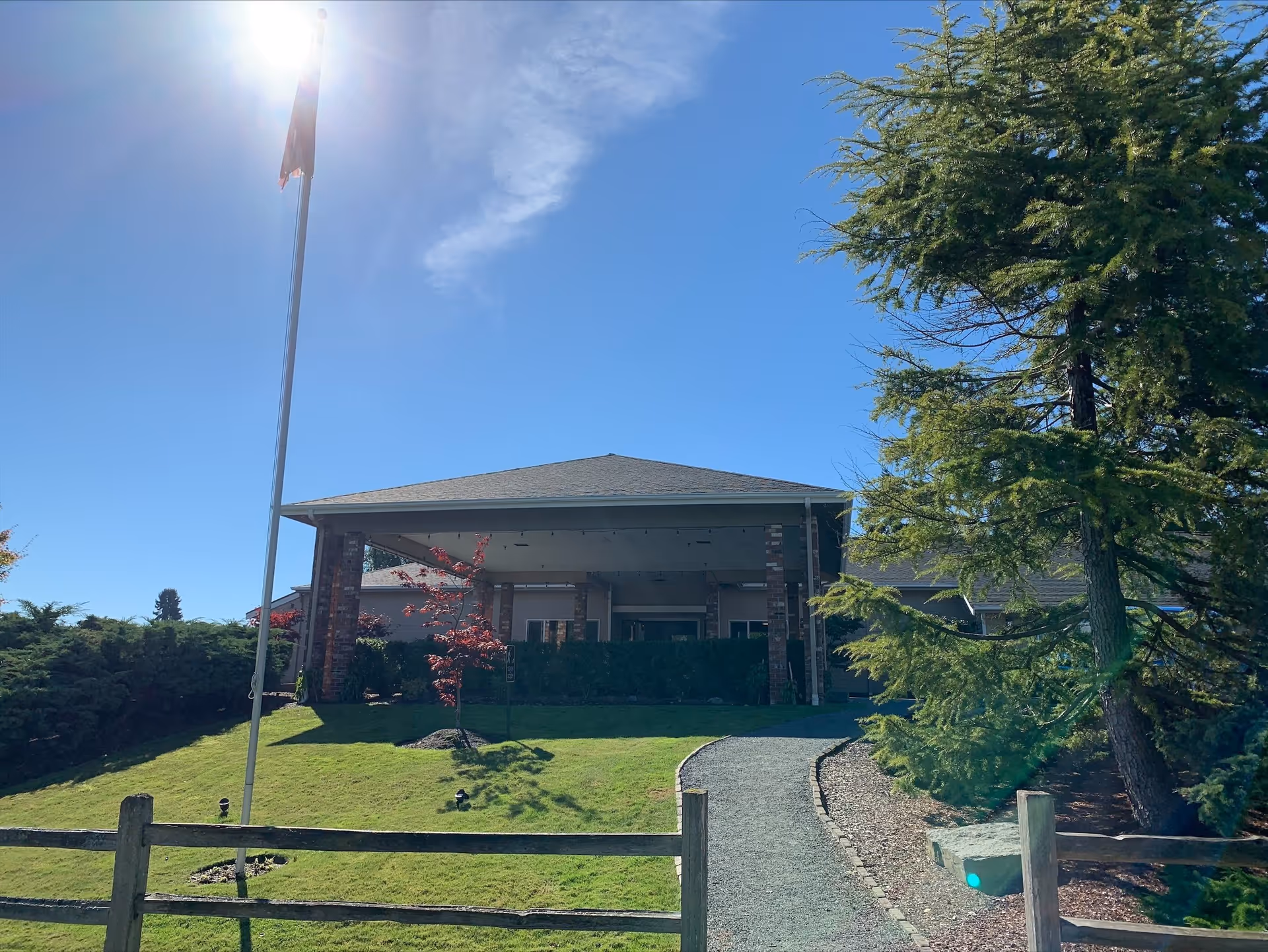Exterior view of Cap Sante Court Retirement Community building with a sloped roof, brick pillars, and a covered entrance. A gravel pathway leads up to the entrance, flanked by green grass, trees, and a wooden fence. The sky is clear and sunny with the sun shining brightly near the top left corner.