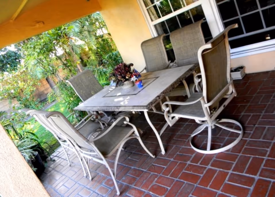 Outdoor patio area with a rectangular table surrounded by six metal chairs with mesh backs and seats. The patio has a tiled floor and is adjacent to a garden with green plants and trees. A window is visible on the wall next to the patio.