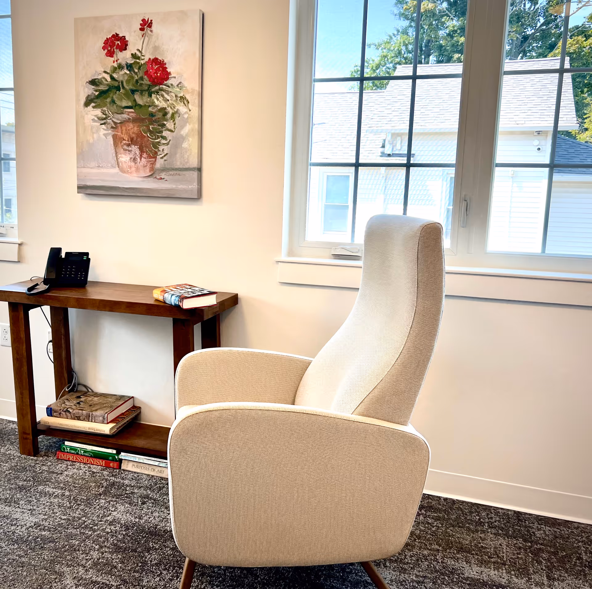A cozy corner of a room featuring a beige upholstered armchair facing a window with white trim. Next to the chair is a wooden console table with a black telephone and several books on top and underneath. A painting of a potted plant with red flowers hangs on the wall above the table. Outside the window, parts of neighboring buildings and green trees are visible.