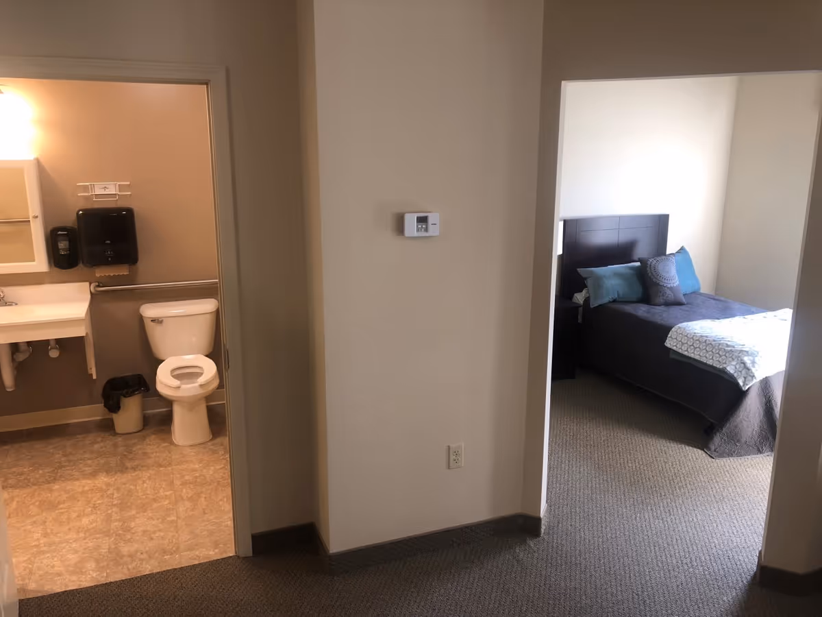 View of a hallway in an assisted living facility showing a bathroom on the left with a toilet, sink, and wall-mounted soap and paper towel dispensers, and a bedroom on the right with a bed made with pillows and a blanket.