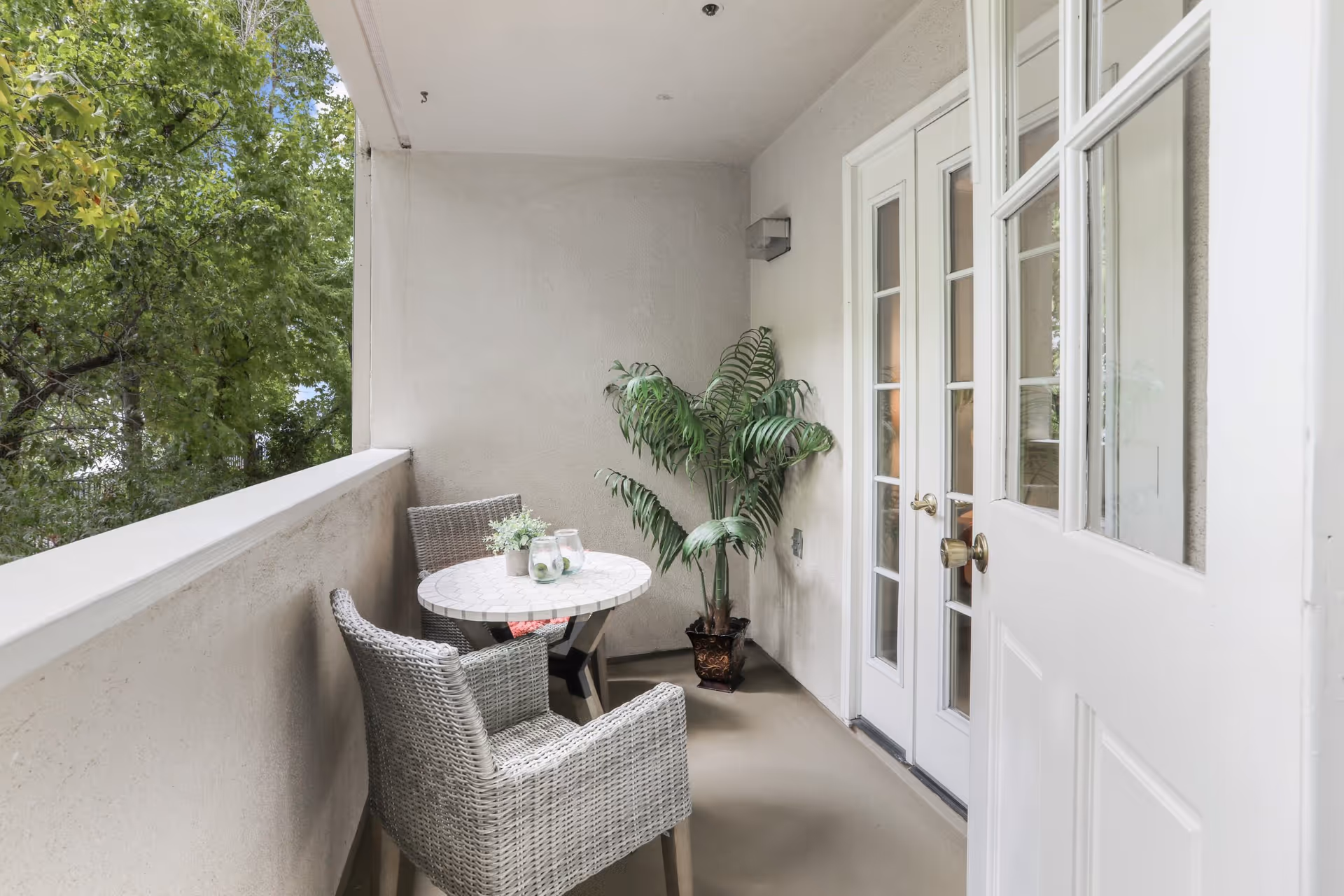 A small outdoor balcony area with two wicker chairs and a round table with a mosaic top. On the table are two glasses and a small potted plant. There is a large potted green plant in the corner near white French doors leading inside. Trees and greenery are visible beyond the balcony railing.
