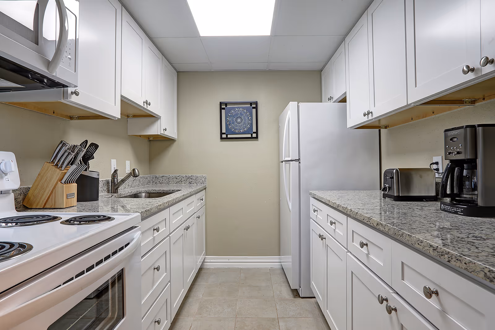 A clean, modern kitchen with white cabinets and granite countertops. The kitchen features a white electric stove, a microwave, a knife block, a sink, a refrigerator, a toaster, and a coffee maker. There is a framed decorative piece on the wall at the end of the kitchen.