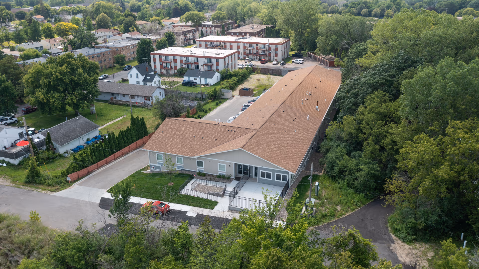 Aerial view of a single-story care facility with a long tan roof, parking area, and surrounding trees and nearby residential buildings.