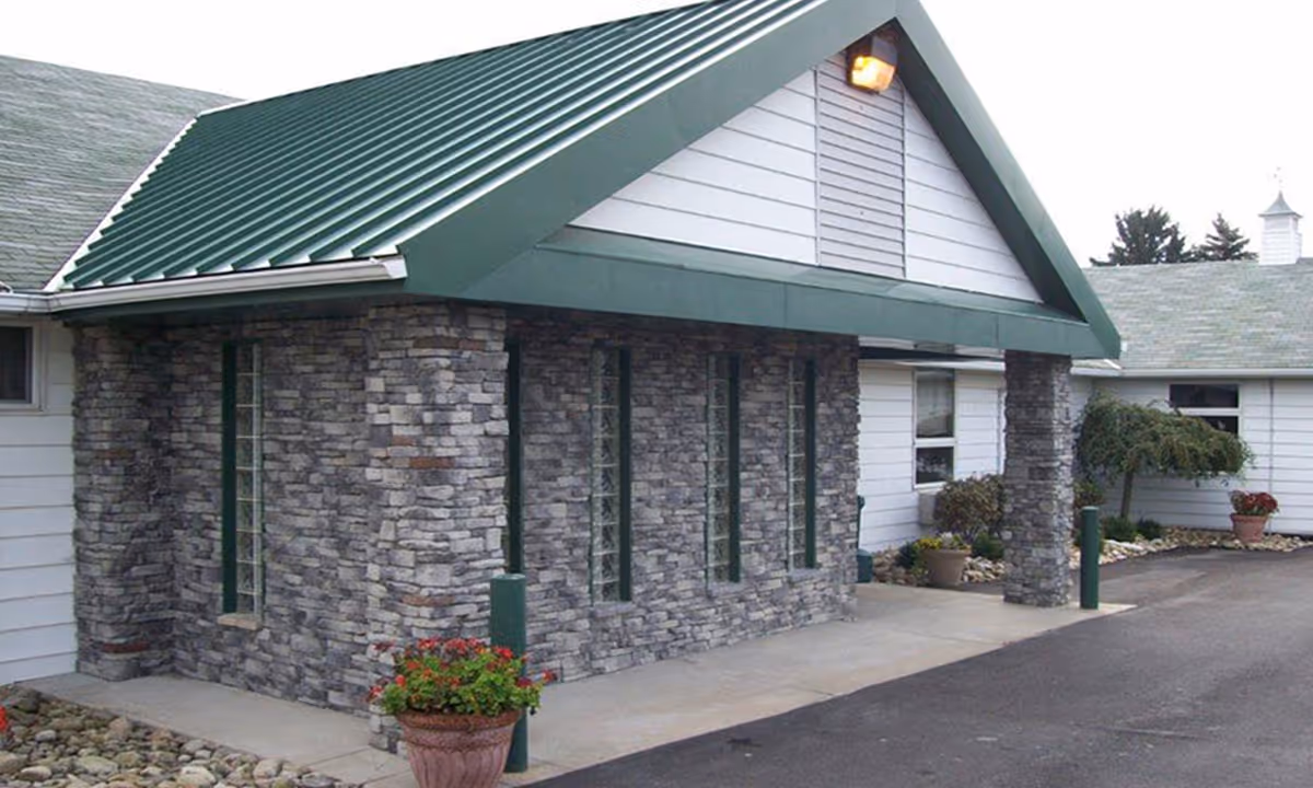 Exterior view of a building with stone and white siding walls, green metal roof, and a covered entrance. There are potted plants and landscaping with rocks around the building.