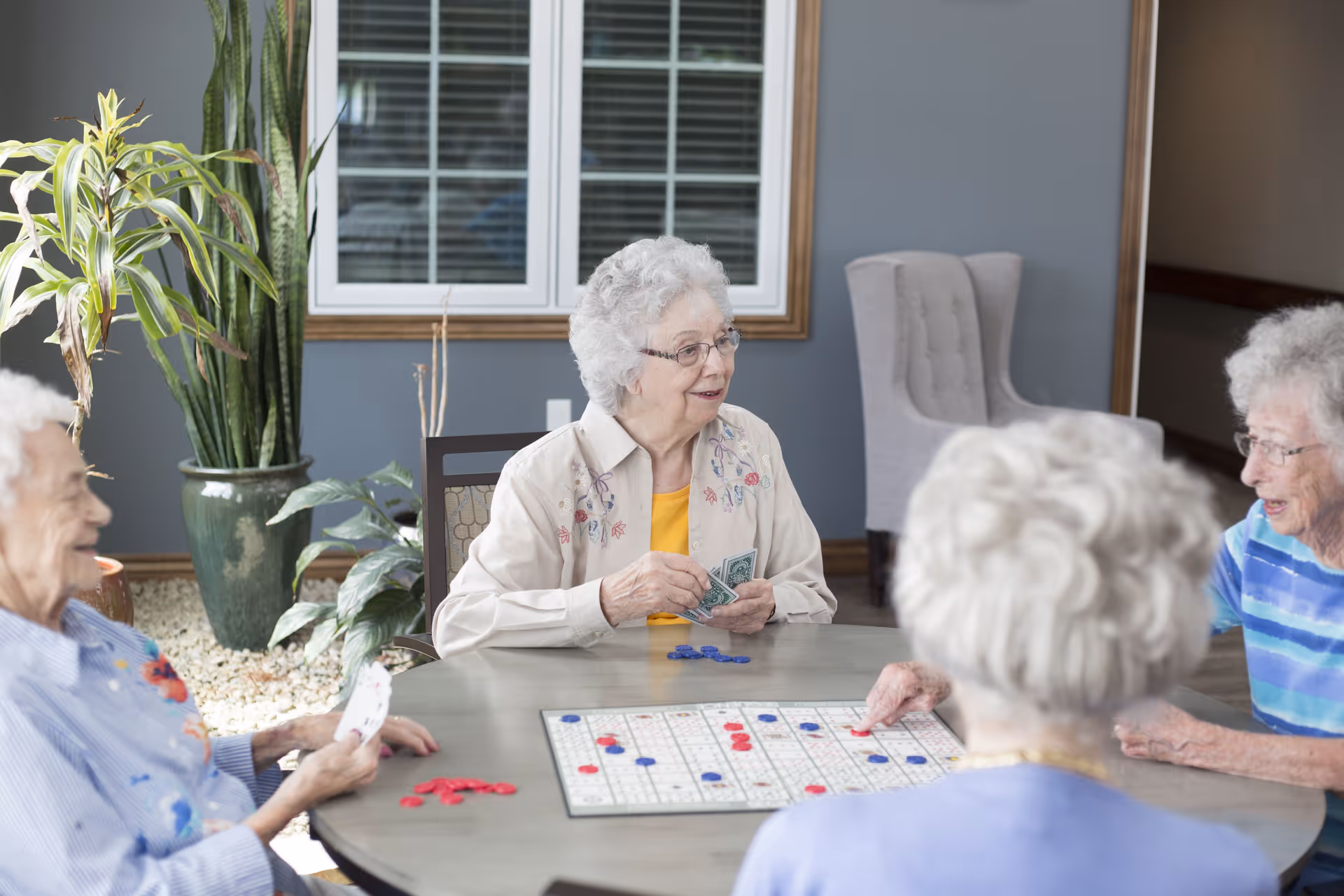Four elderly women sitting around a round table playing a board game with red and blue pieces in a well-lit room with large windows and indoor plants.