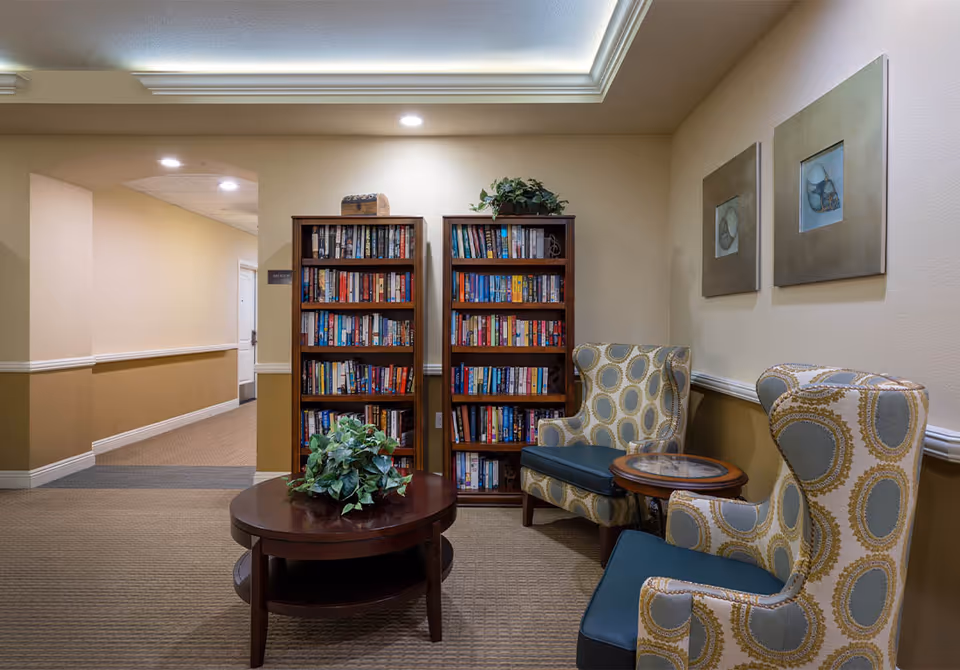 Well-lit reading nook with two patterned armchairs, a round coffee table, and bookshelves against a hallway wall.