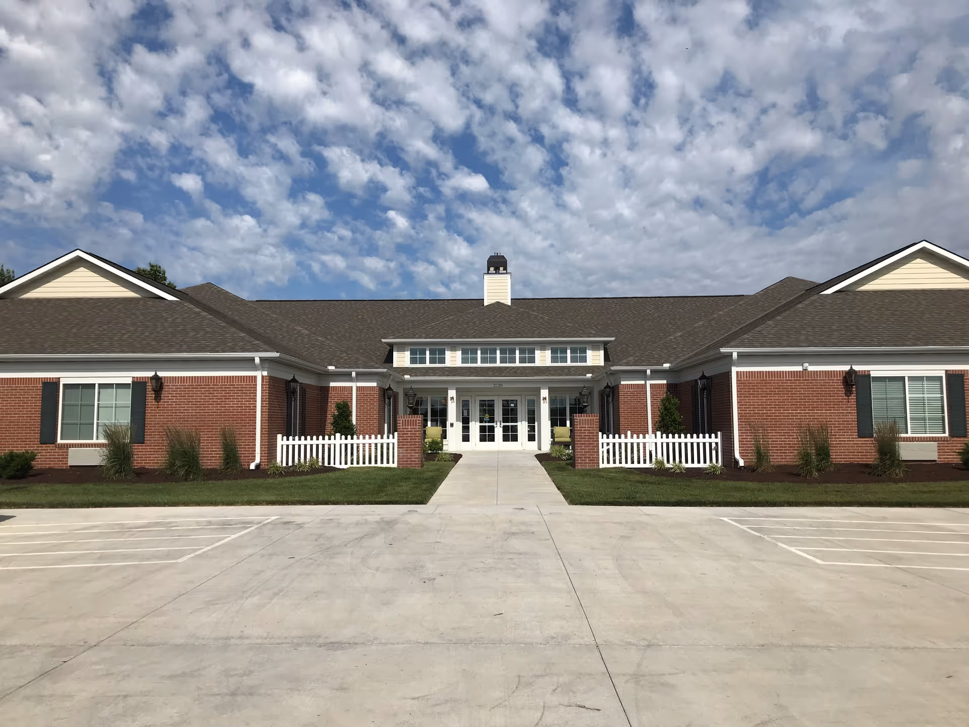 Front exterior view of a single-story brick building with a dark shingled roof, white trim, and a central entrance featuring double glass doors. The building has white picket fences on either side of the entrance and small landscaped areas with shrubs. The sky above is partly cloudy with scattered white clouds.