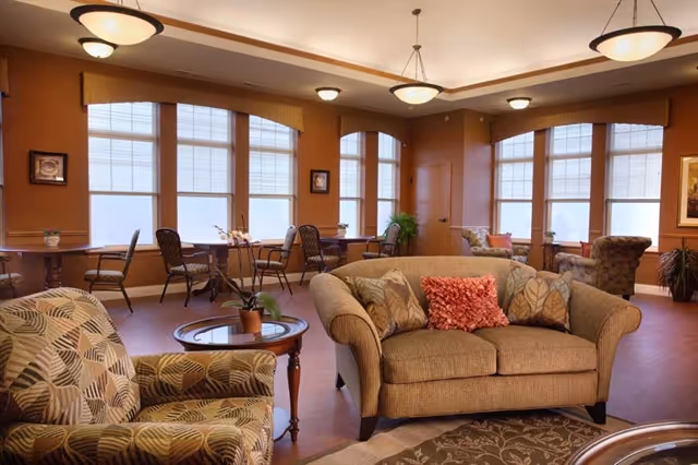 A cozy living room area with a beige sofa adorned with patterned and red throw pillows, a patterned armchair, and a round wooden coffee table with a small plant. The room has large windows with blinds, warm lighting fixtures on the ceiling, and additional seating with tables and chairs near the windows. The walls are painted in a warm brown tone, and there are framed pictures and plants decorating the space.