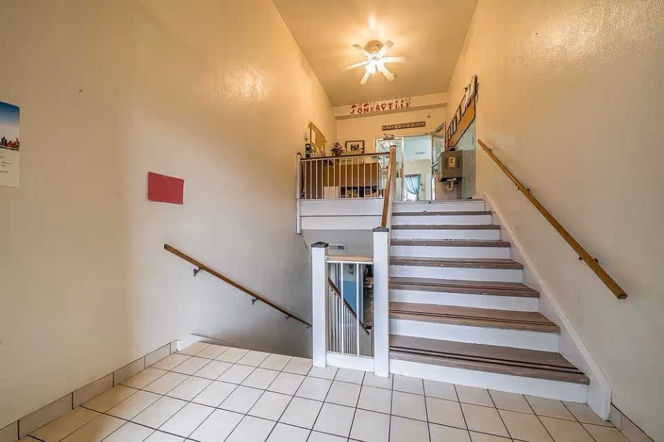 Interior view of a stairwell area in a facility with white tiled floor and beige walls. The stairs have wooden handrails on both sides and lead up to a small landing with a railing. A ceiling fan with a light is mounted above. There are signs and decorations on the wall above the landing, including one that reads 'Jonesville'.