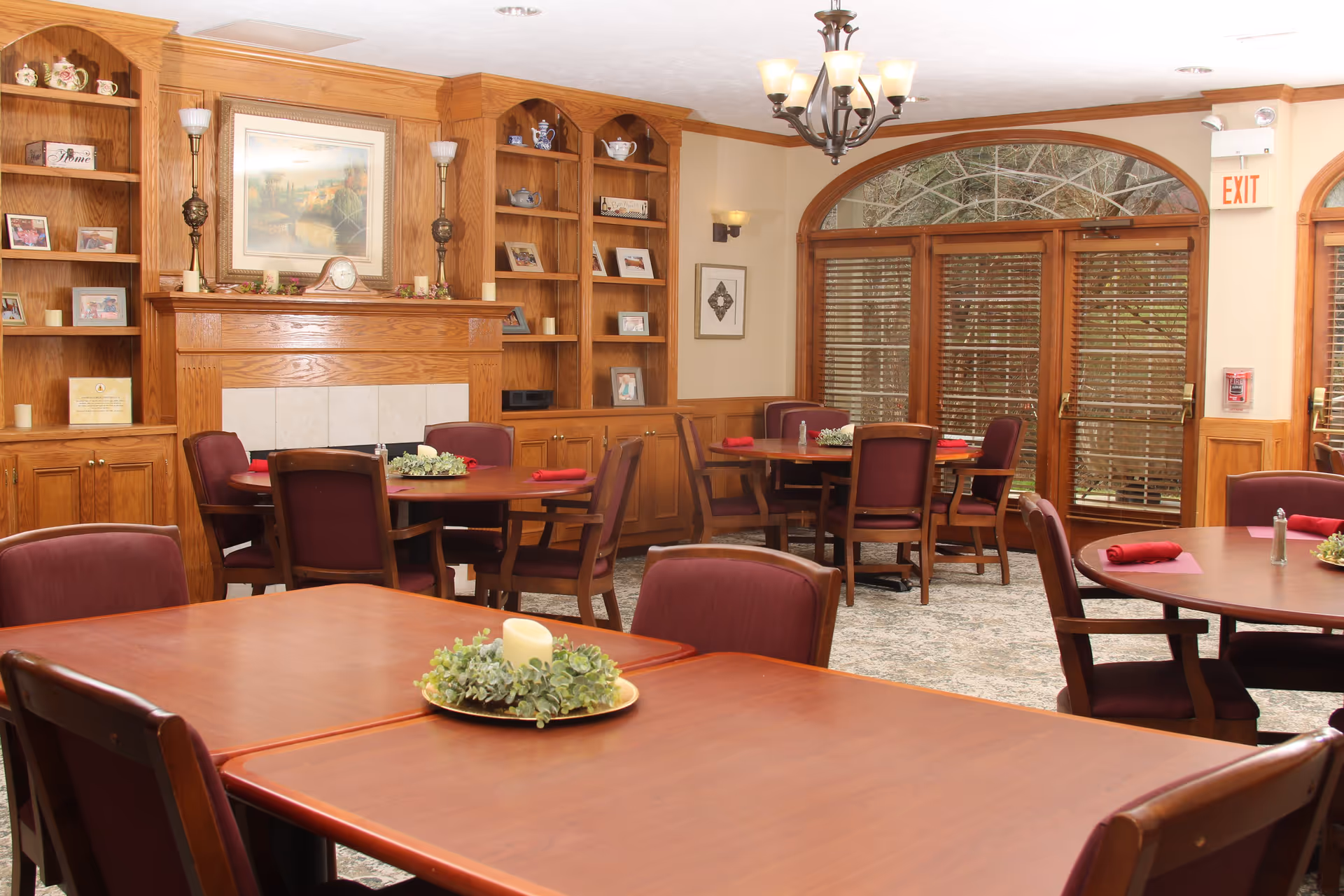 A cozy dining room with wooden tables and maroon cushioned chairs. The room features a wooden fireplace mantel with shelves on either side displaying decorative items and framed photos. Large windows with wooden blinds allow natural light to enter. The tables are set with red napkins and small centerpieces with candles and greenery. A chandelier hangs from the ceiling, and an exit sign is visible above a door.