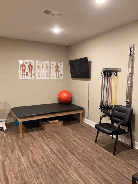 A therapy or exercise room with a black padded treatment table, a red exercise ball, resistance bands hanging on the wall, a black chair, anatomical posters on the wall, and a mounted flat screen TV.